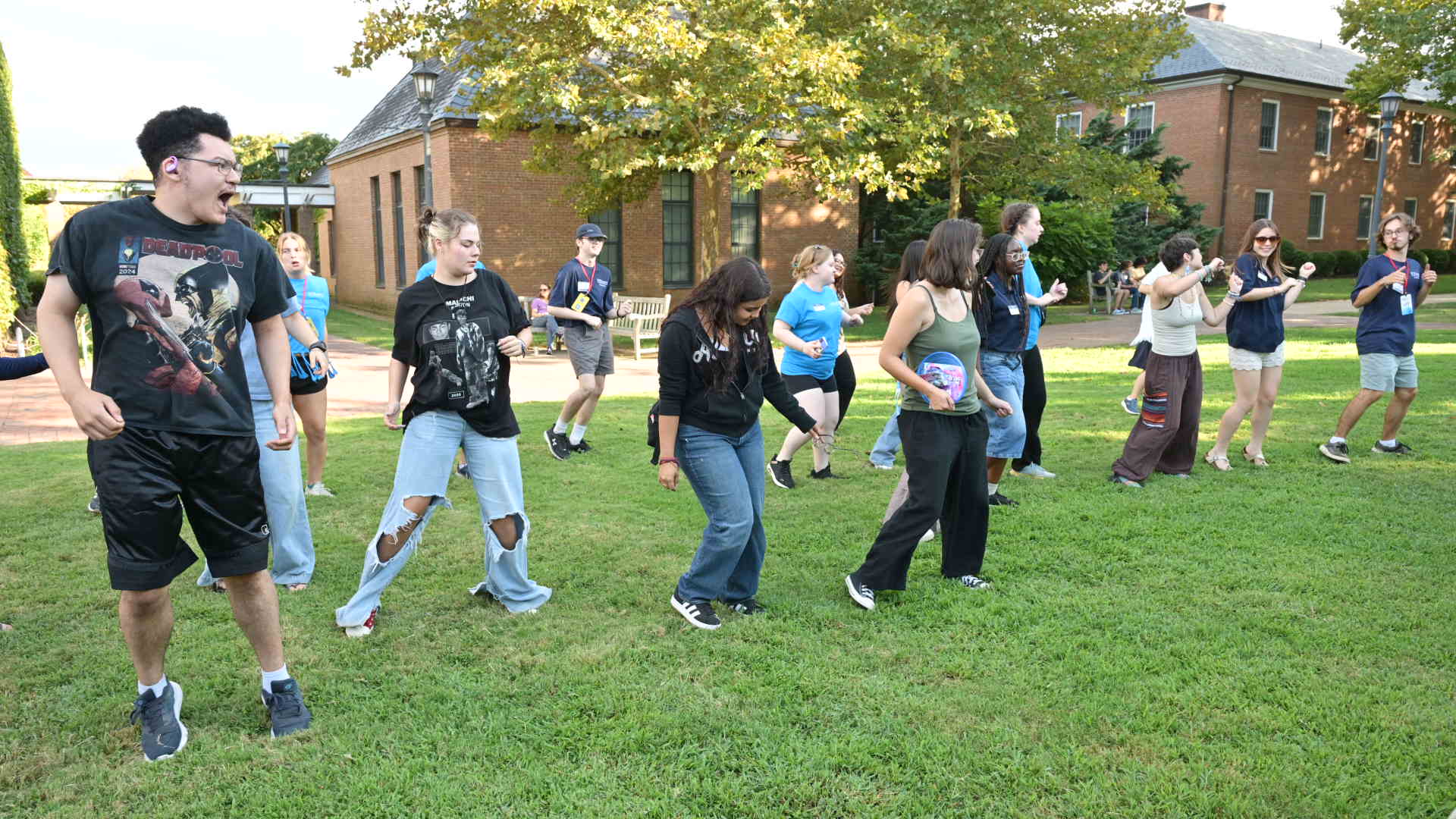 A group of people stand on a lawn, participating in a coordinated outdoor activity or dance, with buildings and trees in the background.