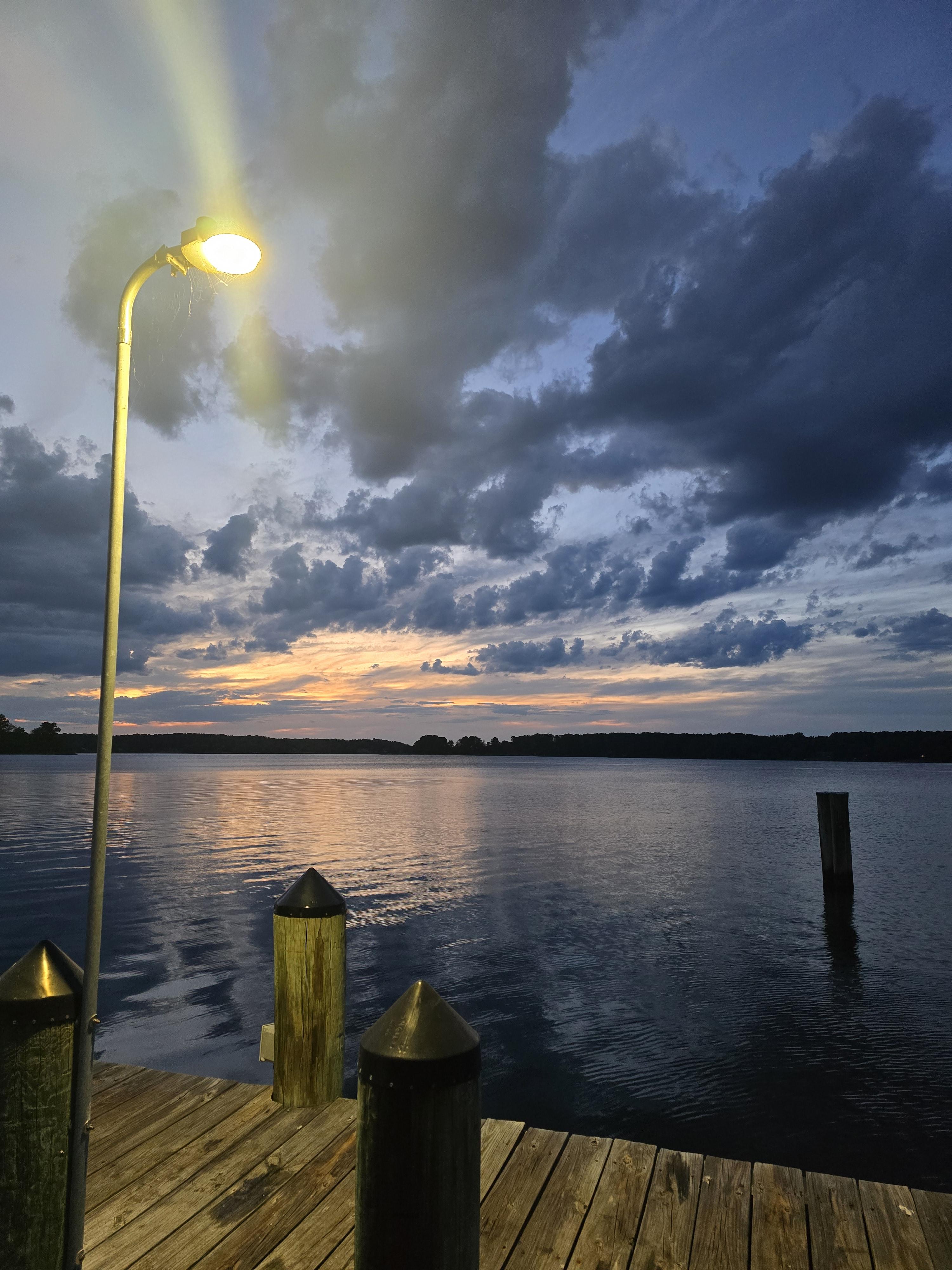 A wooden dock with posts extends over a calm lake at sunset, under a cloudy sky. A tall lamp post is lit on the left, reflecting on the water’s surface.