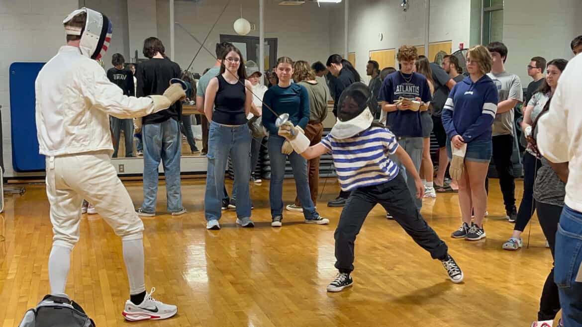 Two people fence in a gym while a group of onlookers watch; one fencer is lunging, and the other stands ready.