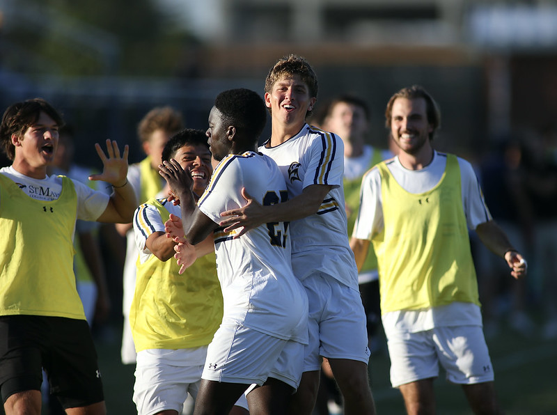 Soccer players in white uniforms celebrate together on the field, with teammates in yellow pinnies joining in and smiling in the background.