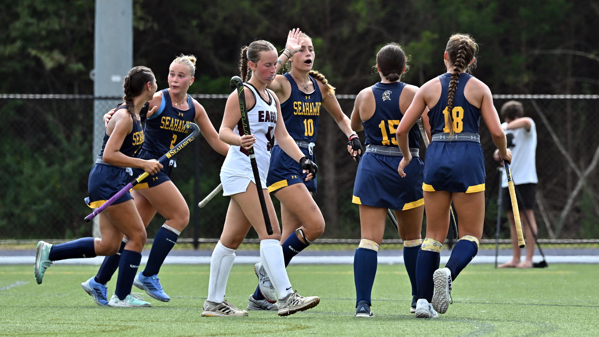 A field hockey team in navy and yellow uniforms celebrates on the field as two players in white walk away, with trees and a fence in the background.