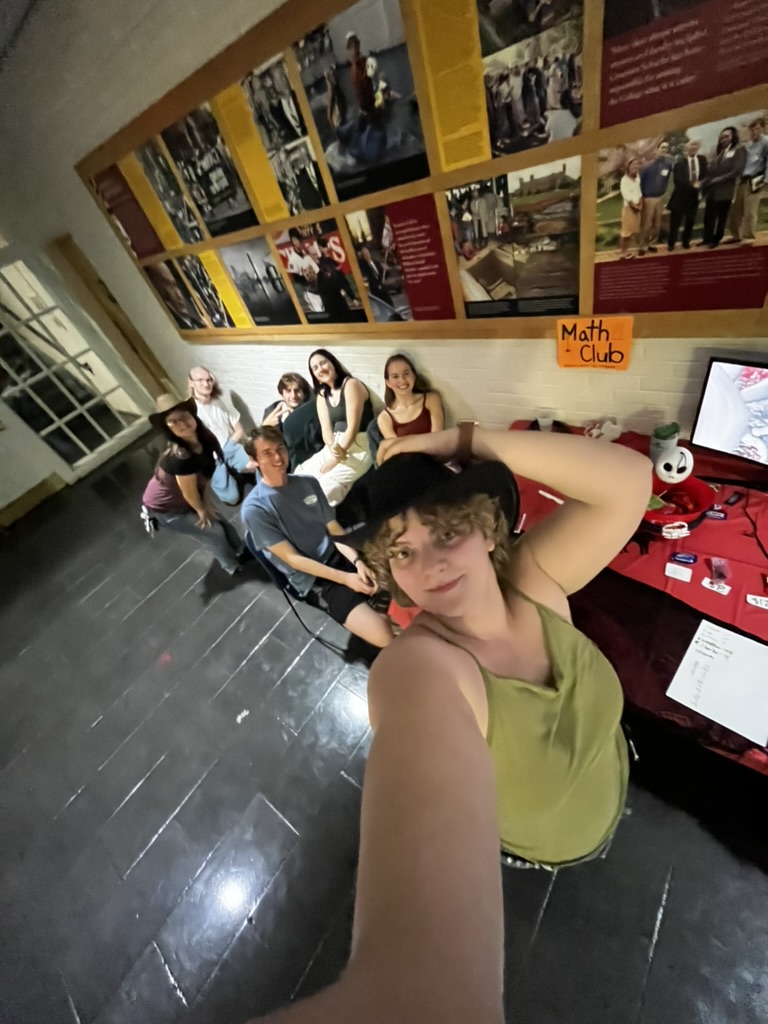 Seven people pose for a selfie in front of a table labeled "Math Club," with photos and posters displayed on the wall behind them.