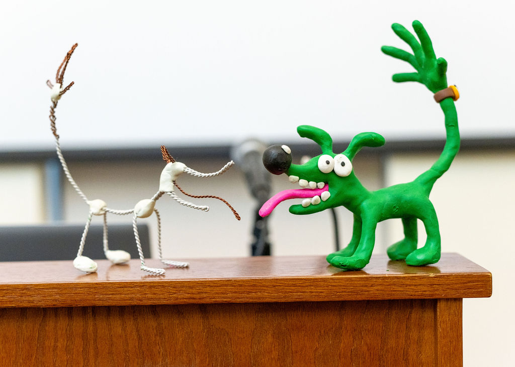 Two dog sculptures, one made of wire and white beads, the other made of green clay with exaggerated features, are displayed on a wooden surface.