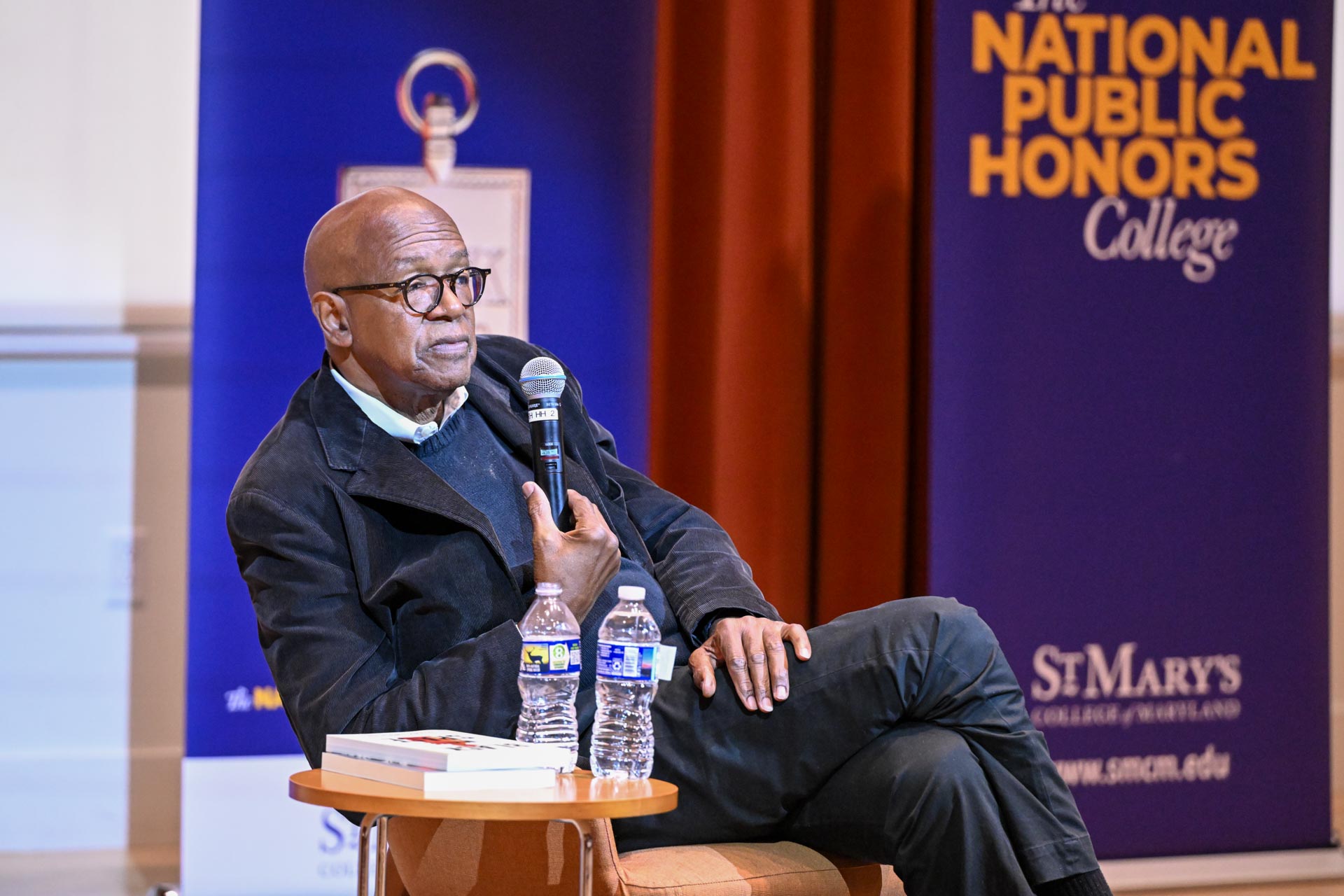 A man sits on stage holding a microphone, with two water bottles and a book on a table beside him. A "National Public Honors College" banner is in the background.