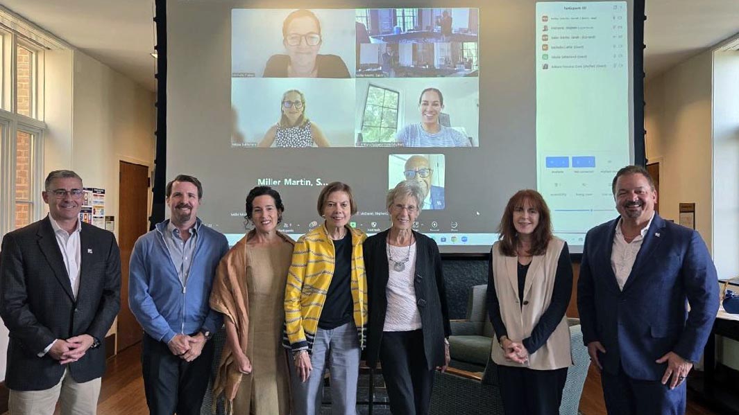 Eight people stand in a row indoors, with a large screen behind them showing a video call with five additional people.