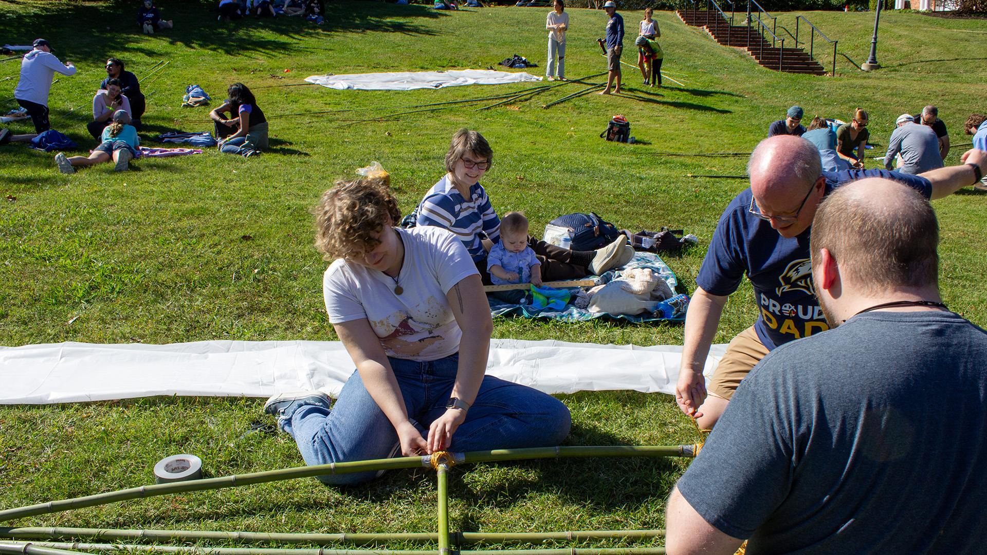 Several people sit on the grass assembling a bamboo structure, while others relax on blankets in the background on a sunny day.