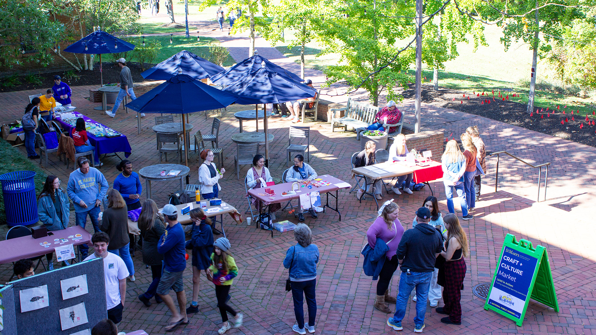 Outdoor campus event with several tables set up under umbrellas, people walking and interacting, and a green sign advertising a culture fair.