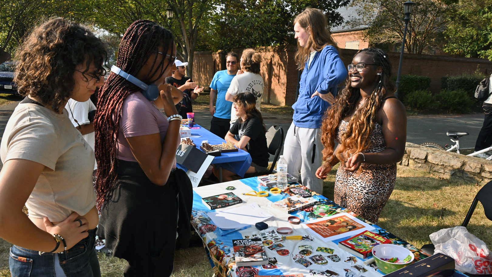 People interact at an outdoor table covered with colorful artwork and crafts during a community or school event on a sunny day.