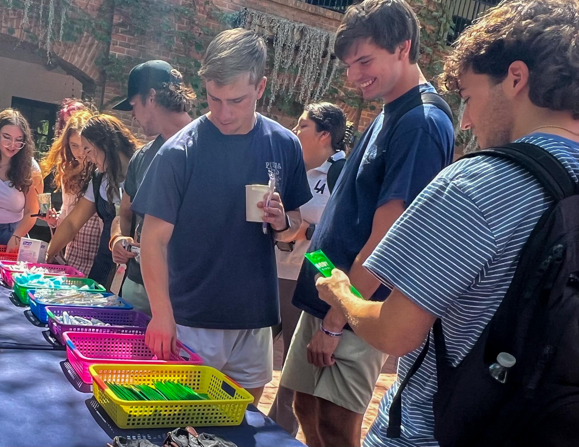 A group of young adults browse colorful baskets of items on a table at an outdoor event. One person holds a drink, another examines a green object.