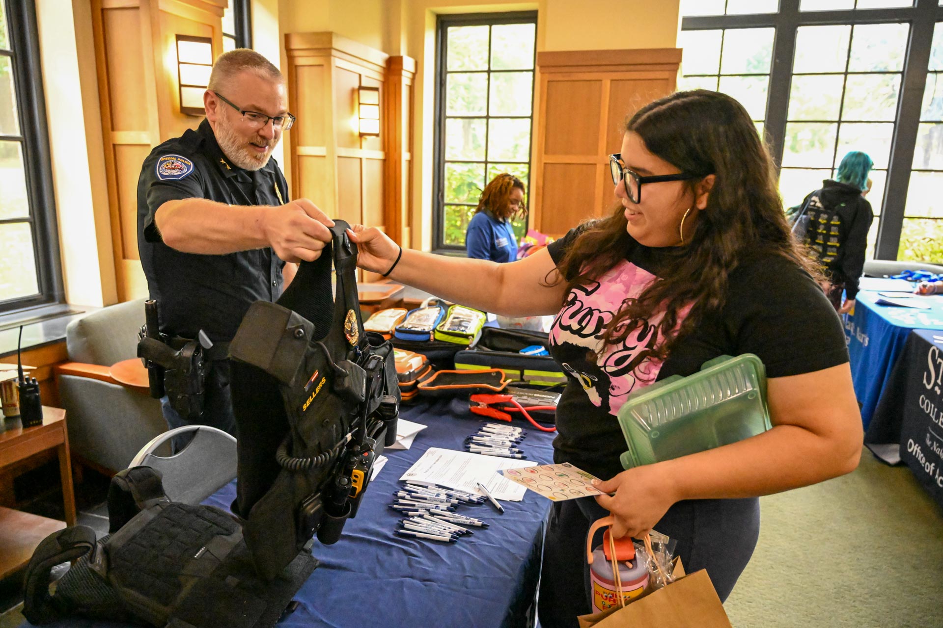 A police officer shows a tactical vest to a woman at an indoor event table with informational materials and giveaway items.