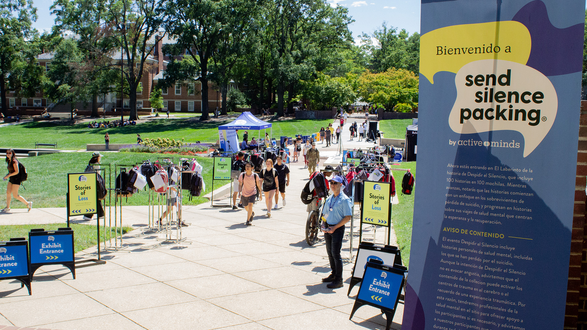People walk through a campus exhibit displaying backpacks and informational signs for the "Send Silence Packing" mental health awareness event.
