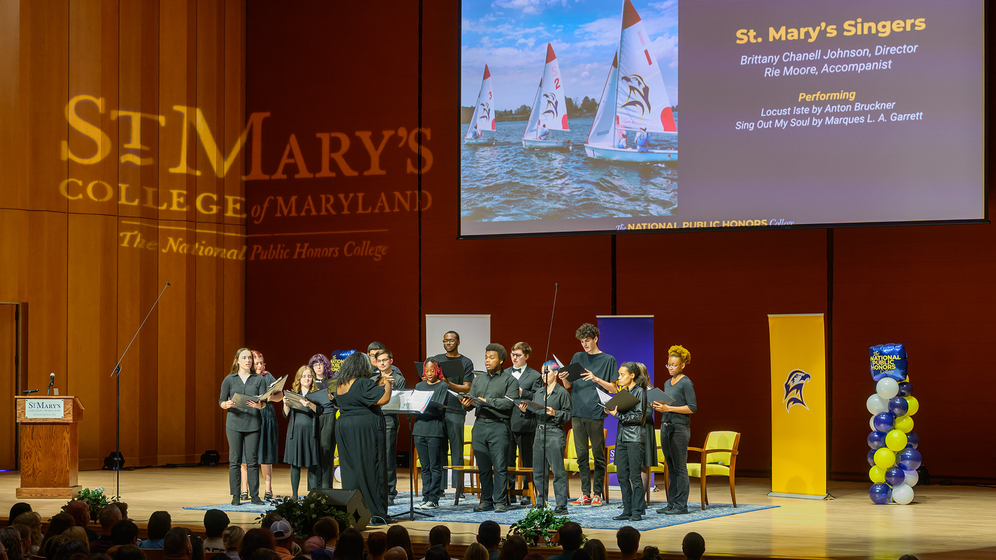 A choir performs on stage at St. Mary’s College of Maryland, with a projected presentation and audience seated in the foreground.