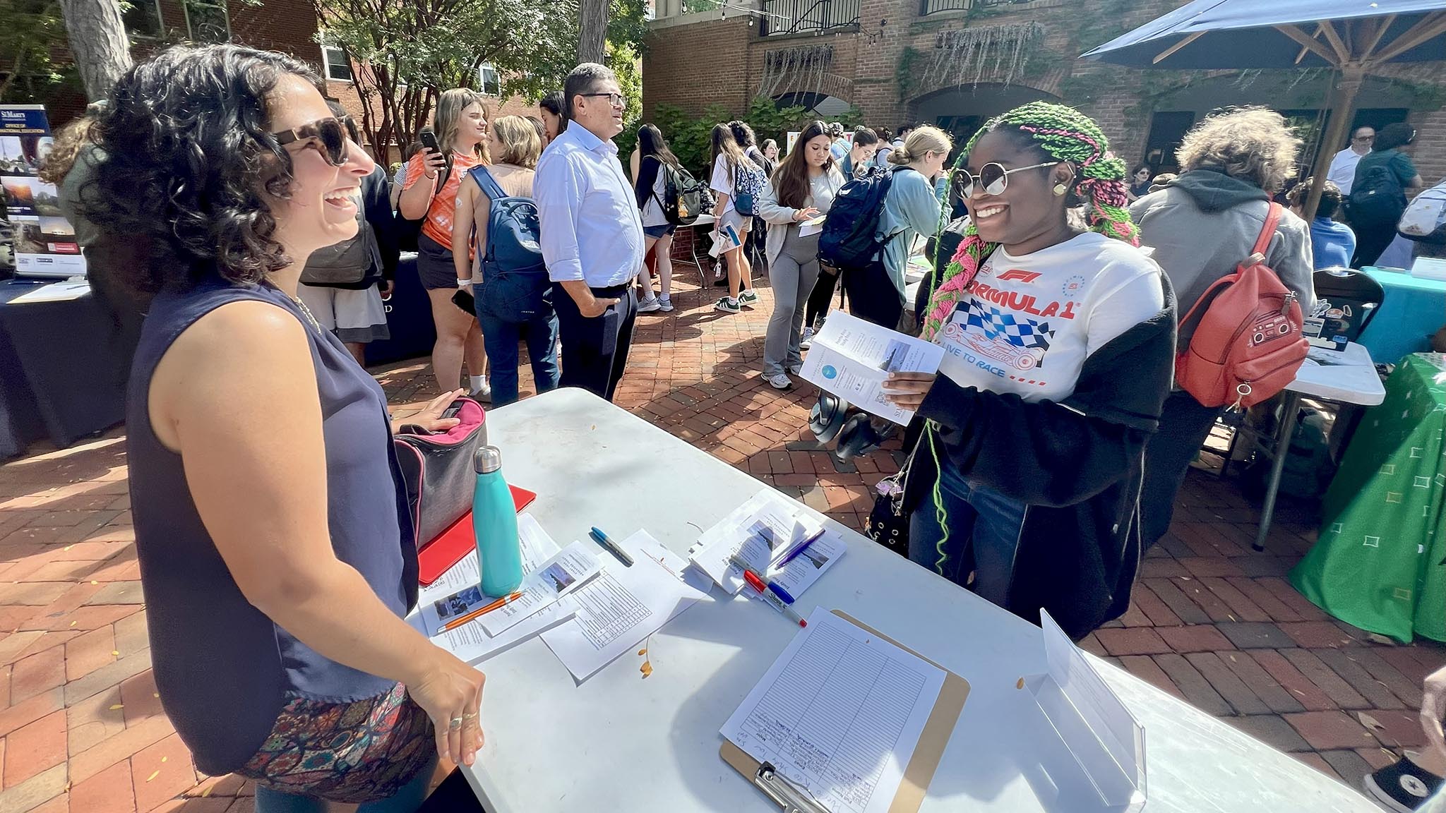 Two women stand at a table with papers and pens, talking and smiling, while other people gather at an outdoor event with booths in the background.