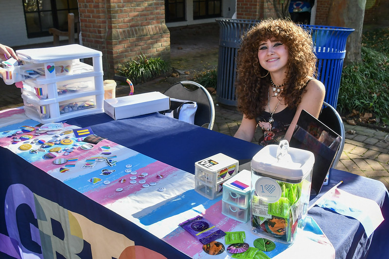 A person with curly hair sits at a table covered with pride-themed pins, stickers, and buttons, outdoors in a sunny area, behind a "GR" banner.