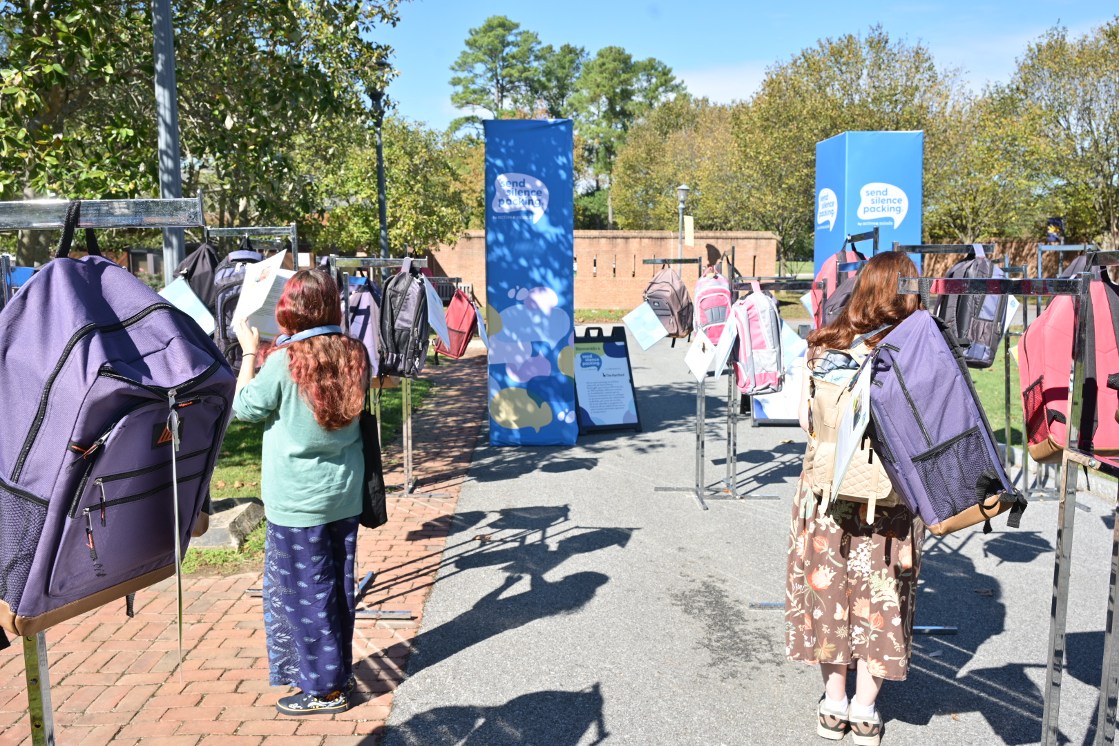 Two people stand among rows of large backpacks displayed outdoors on metal racks, with informational signs and trees in the background.