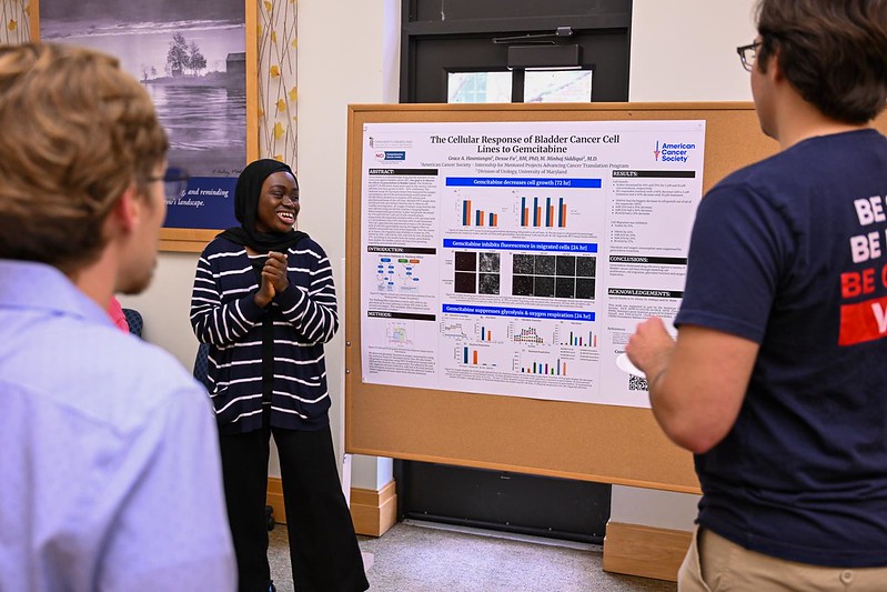 A woman presents scientific research on cancer cell response to three people in front of a poster display in a bright room.