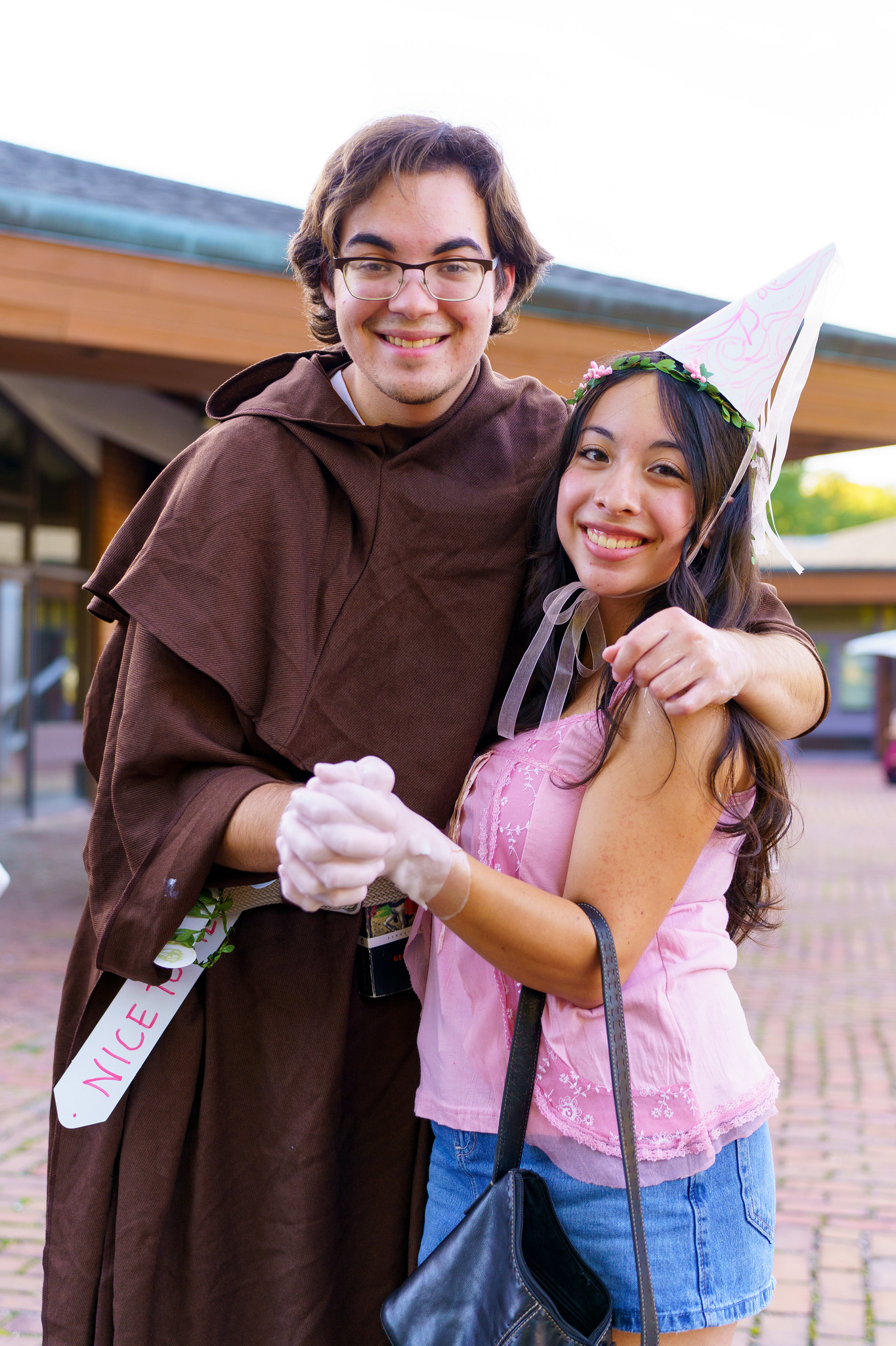 Two people pose outdoors; one wears a brown robe and sash, the other wears a party hat, pink top, and carries a black purse. They are smiling and holding hands.