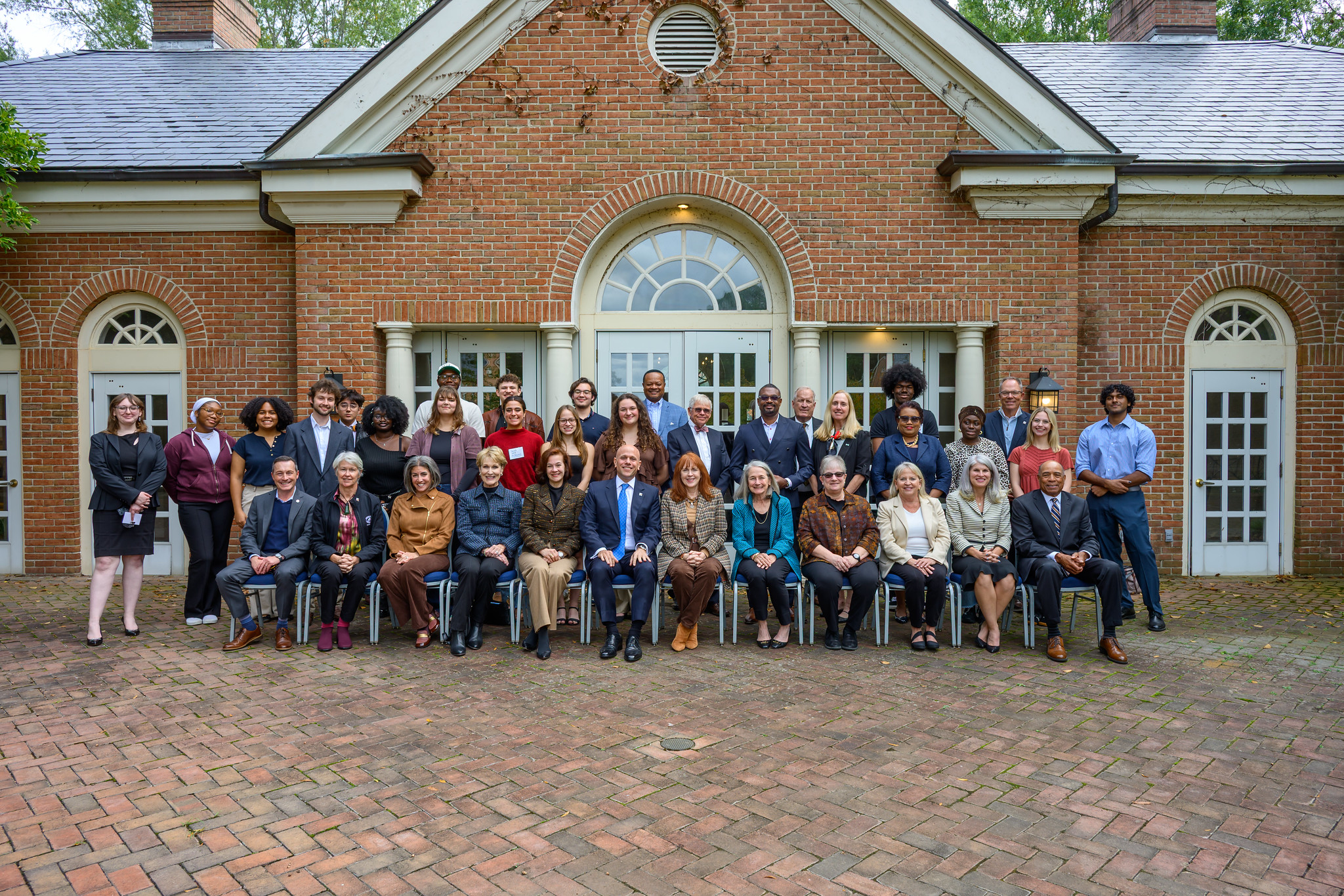 A group of people pose for a formal photo in front of a brick building with arched windows and doors.