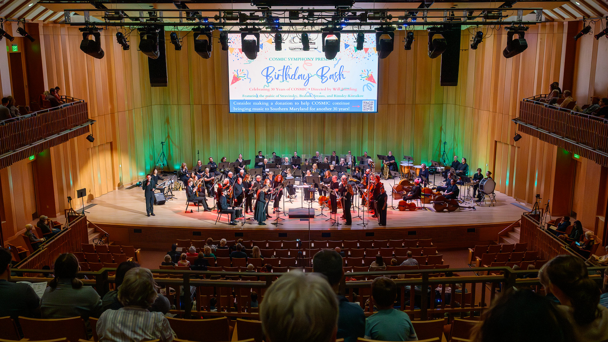 A symphony orchestra performs on a well-lit stage in a concert hall, with musicians seated and some standing; audience members watch from the seats.