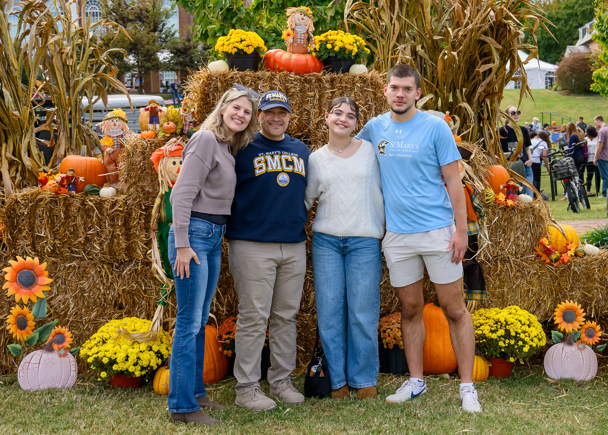 Four people pose in front of a fall-themed display with hay bales, pumpkins, sunflowers, and autumn decorations outdoors.