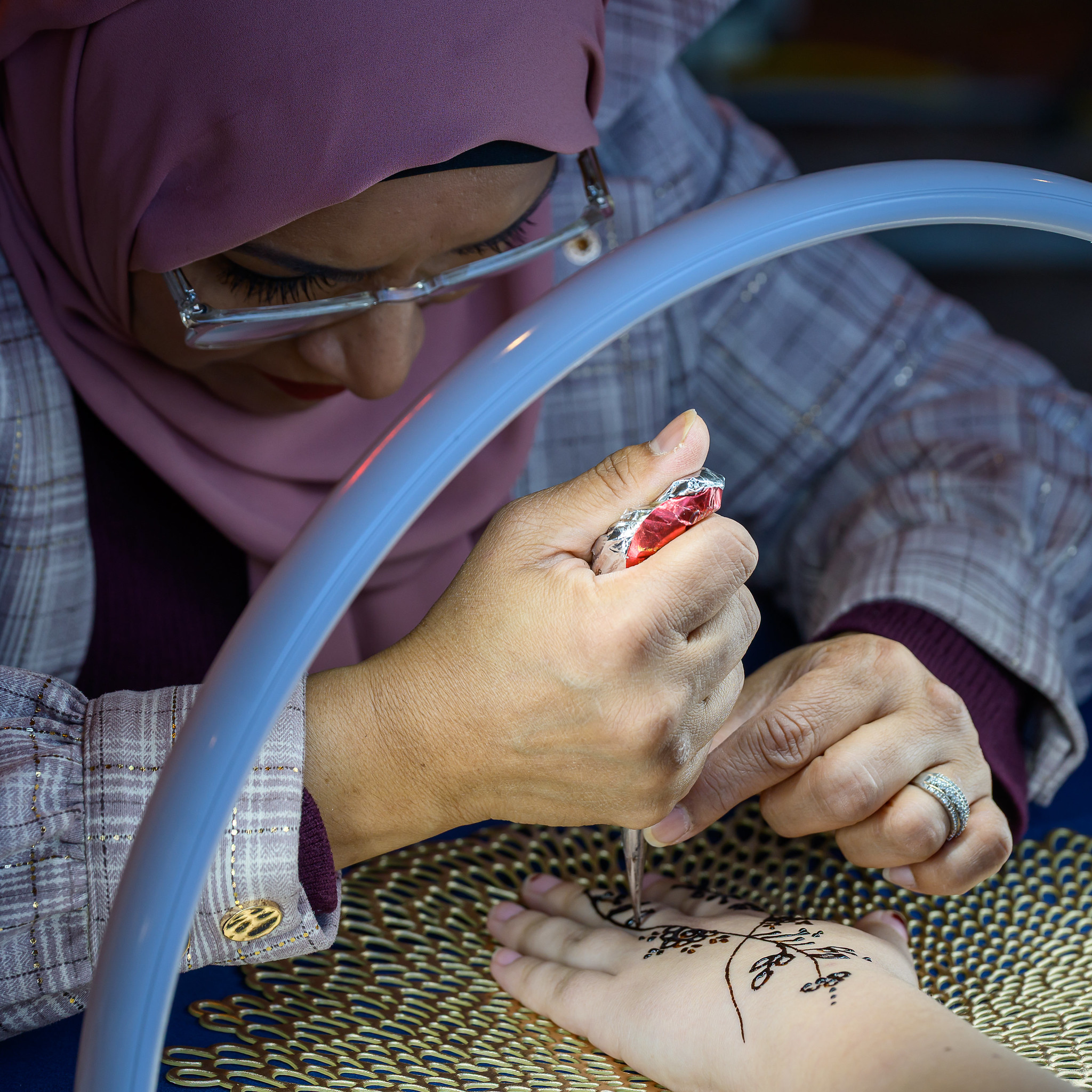 A person in a hijab applies intricate henna designs on another person's hand under a bright ring light.