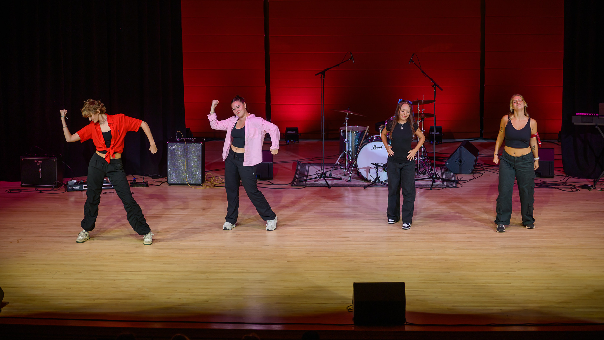 Four people perform a dance routine on stage in front of musical instruments and microphones, with red lighting in the background.