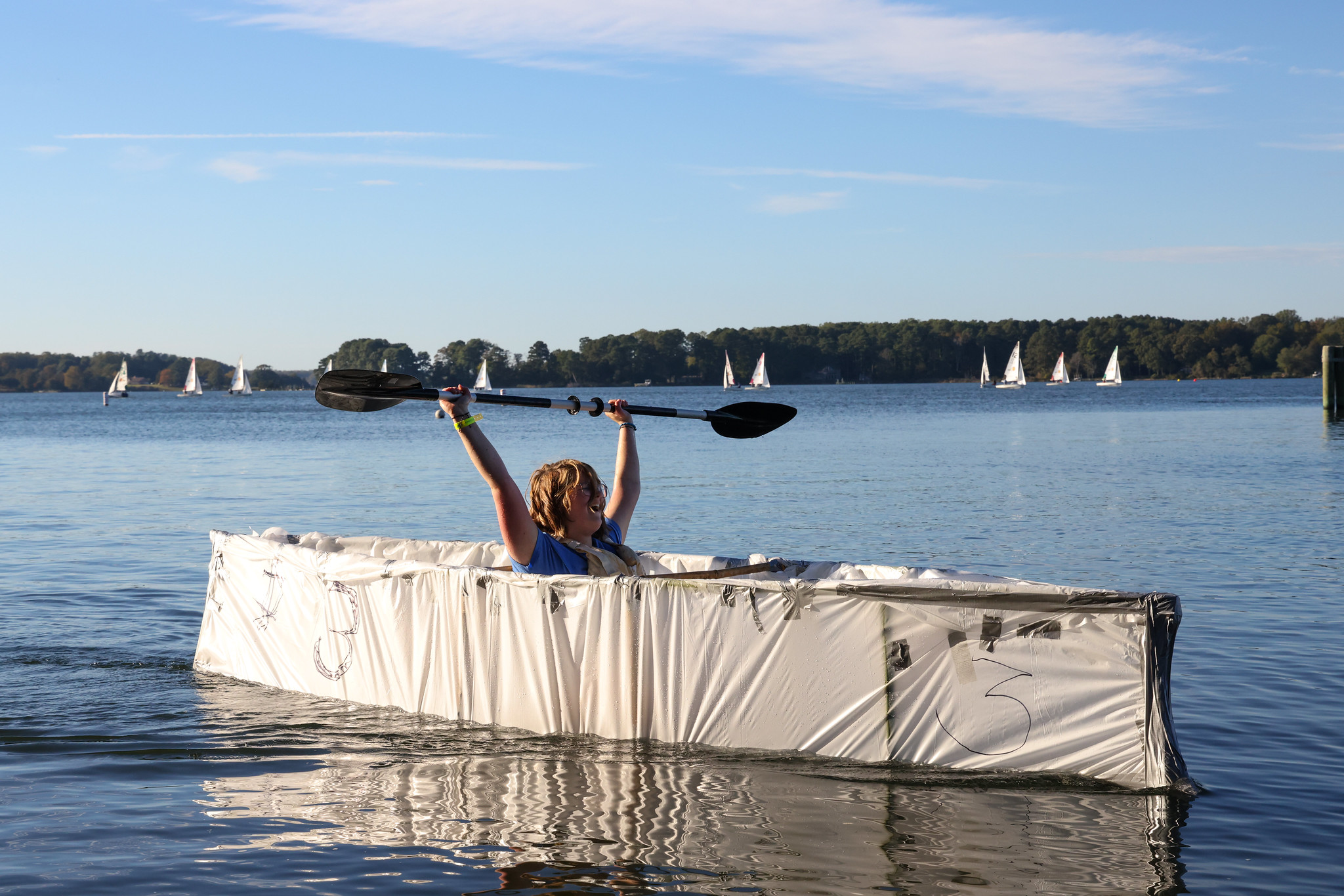 A person sits in a homemade cardboard boat on a lake, raising a paddle overhead. Sailboats are visible in the background under a clear sky.
