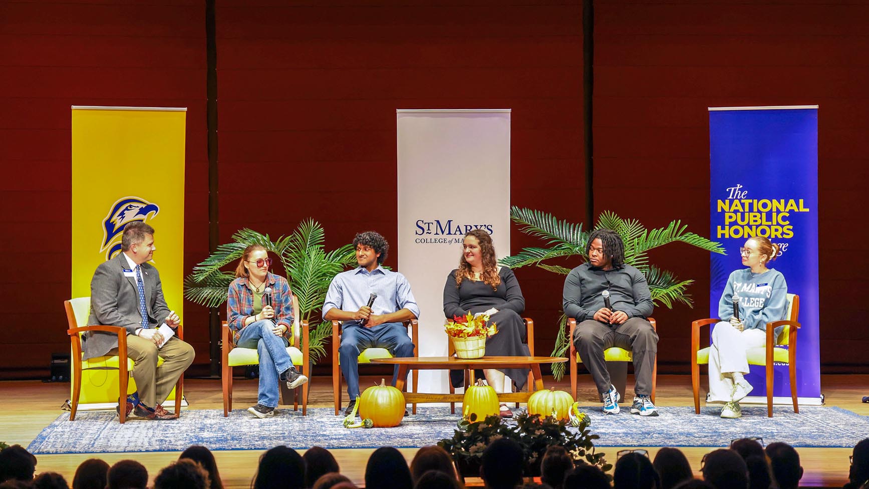 Six people sit on stage in chairs during a panel discussion, with banners, plants, and pumpkins as decoration and an audience in the foreground.