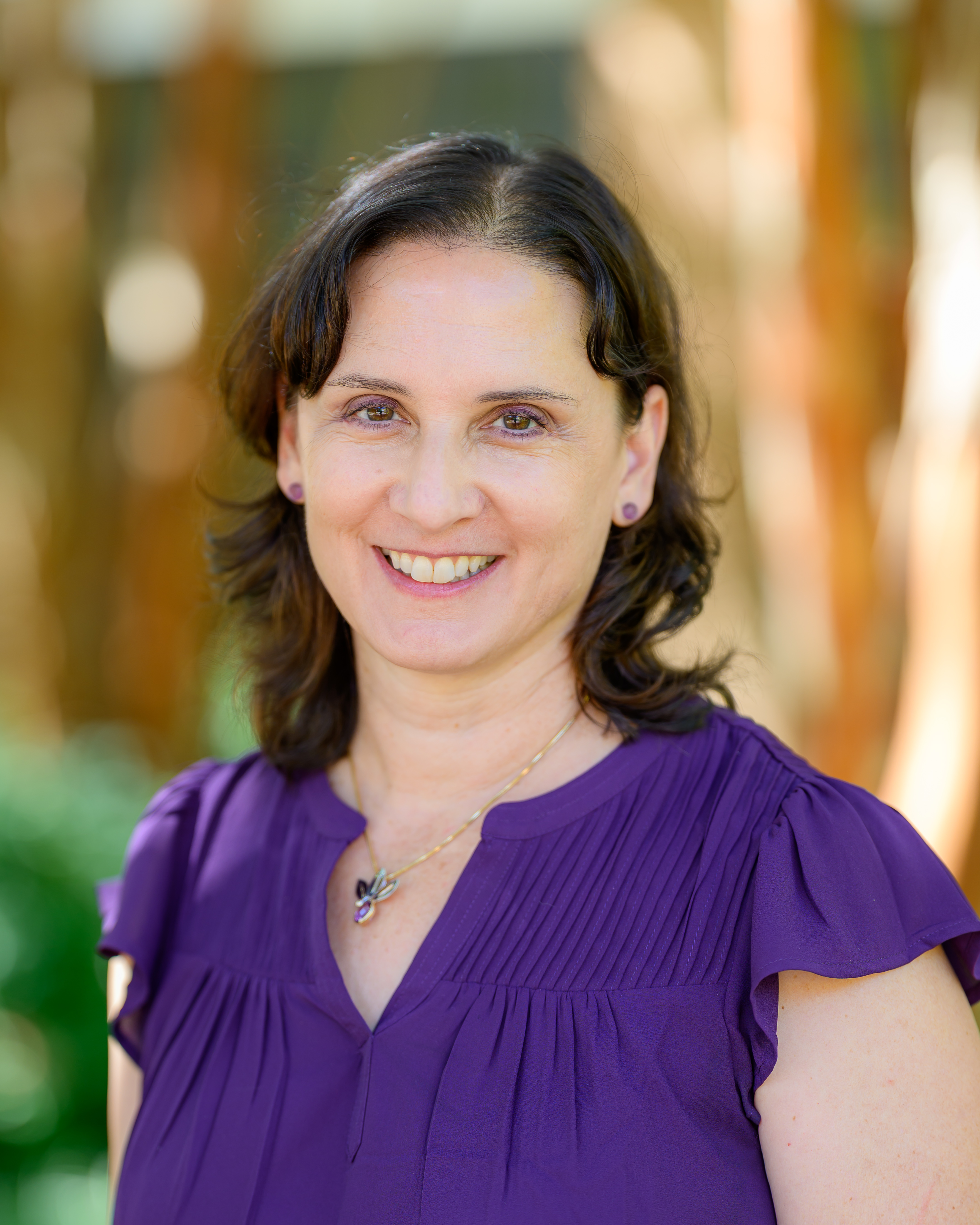 A woman with shoulder-length brown hair wearing a purple blouse and necklace stands outdoors, smiling at the camera.