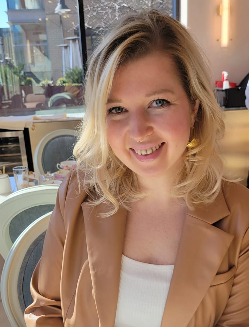 A woman with blonde hair, wearing a tan blazer and white top, smiles while sitting at a table in a sunlit café or restaurant.