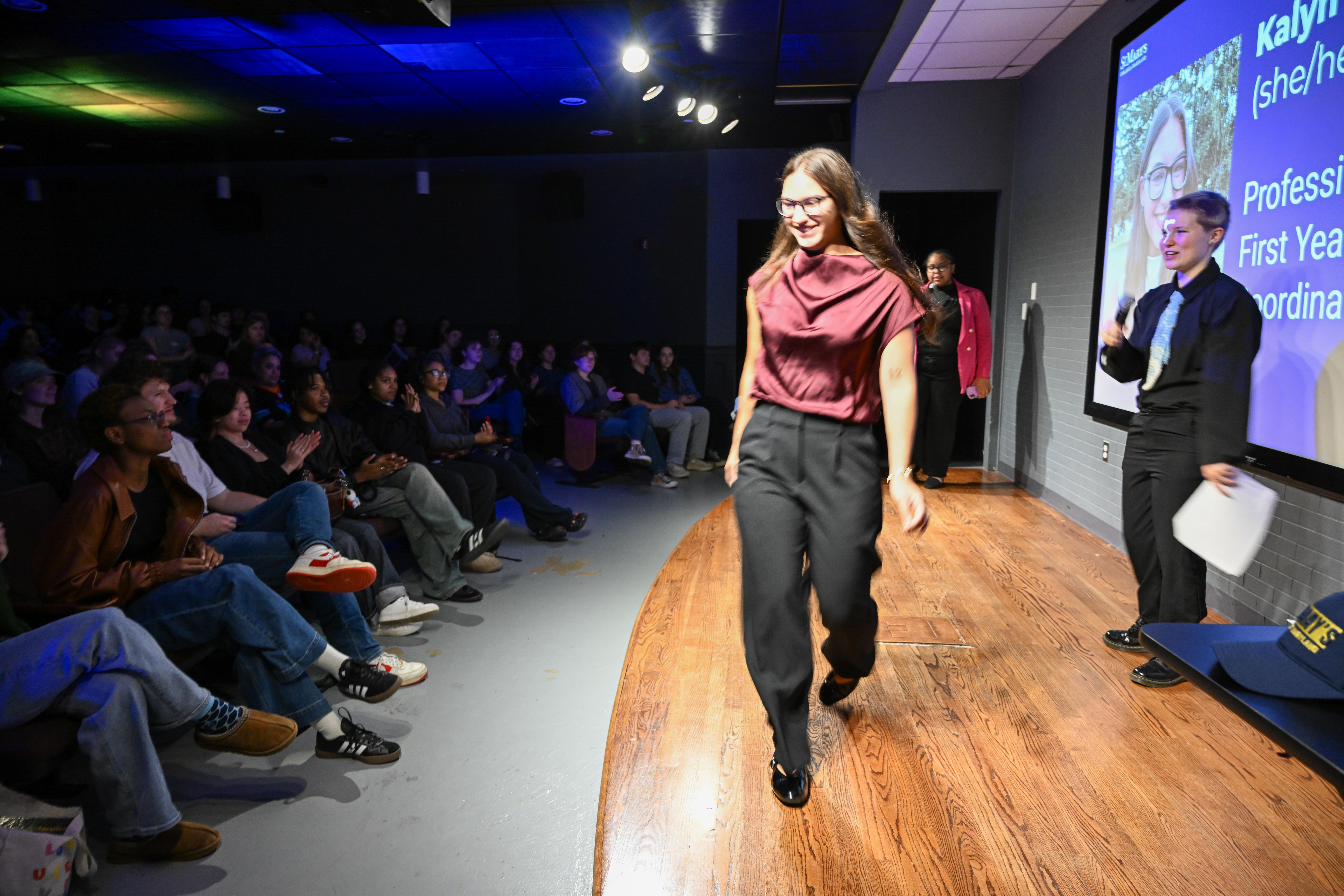 A person walks across a stage in front of an audience, while two people stand by a screen displaying a professional profile and photo.