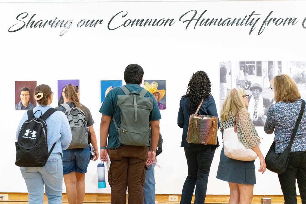 A group of people with backpacks and bags view artwork on a gallery wall beneath a sign that reads "Sharing our Common Humanity.