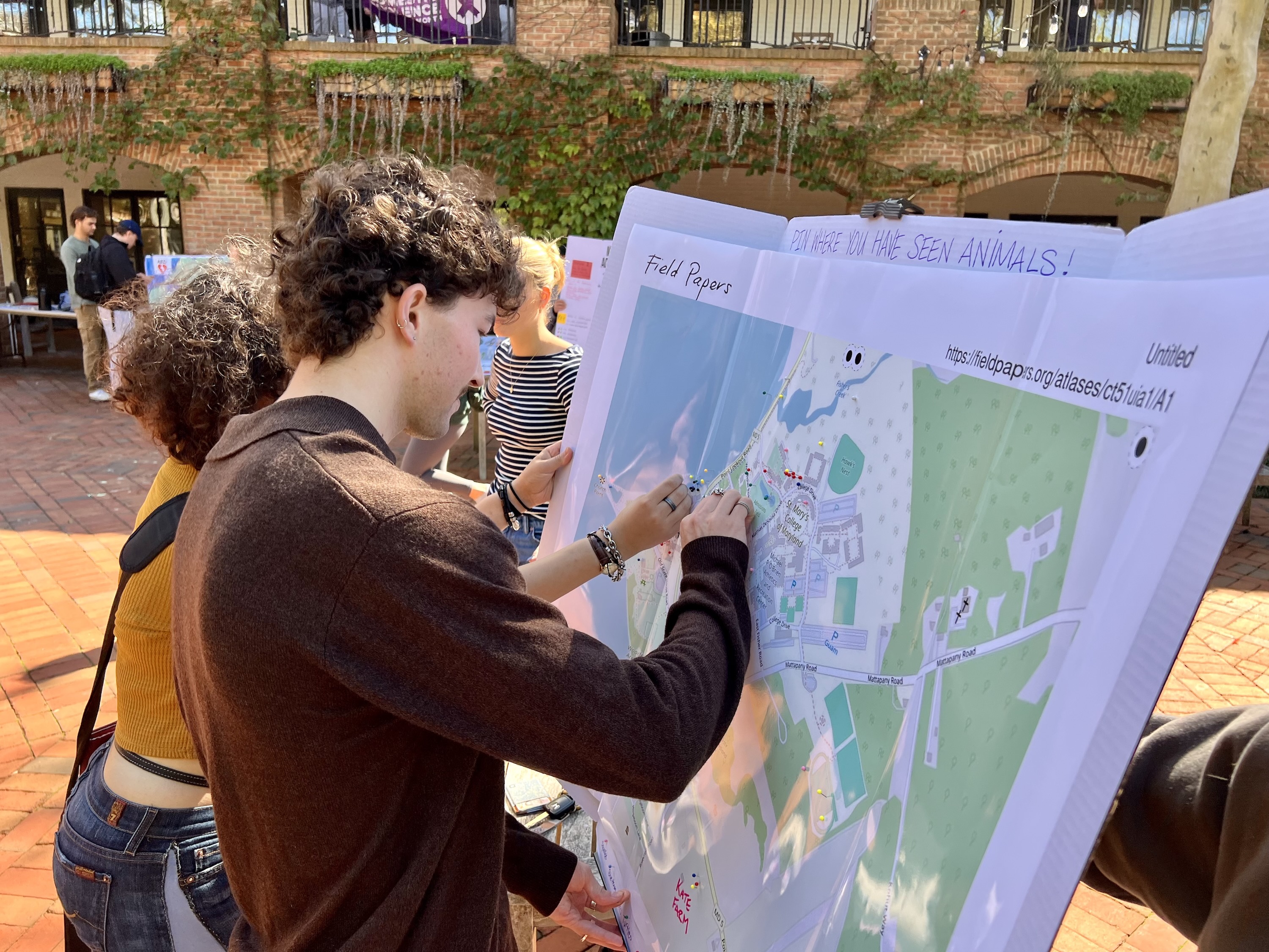 People stand outdoors examining and marking a large printed map spread out on a table in a sunny courtyard setting.