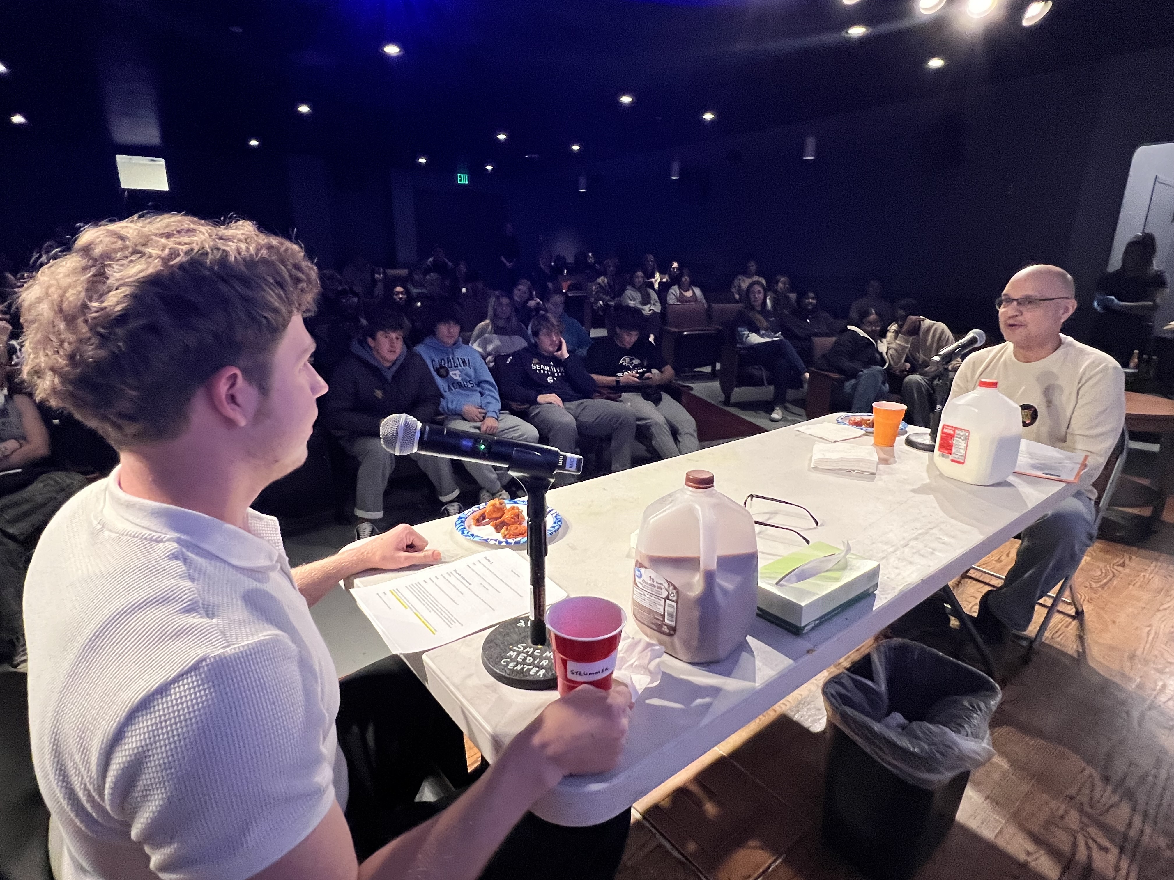 Two people sit at a table on stage facing an audience, with a microphone, drinks, milk jugs, snacks, and napkins on the table under bright lights.