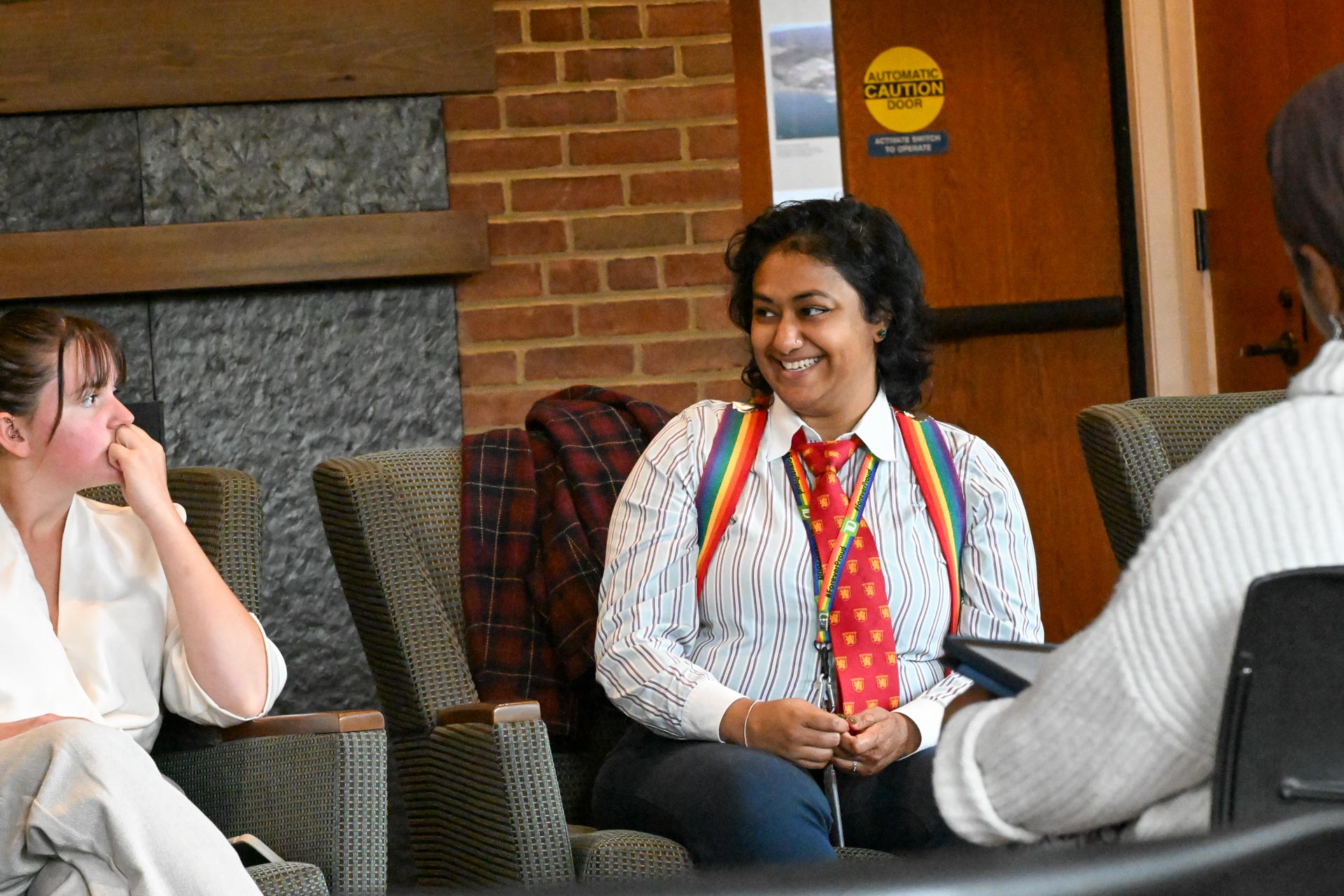 Three people sit in chairs having a conversation; one person in the center wears a striped shirt, rainbow suspenders, and a red tie, smiling at another person.