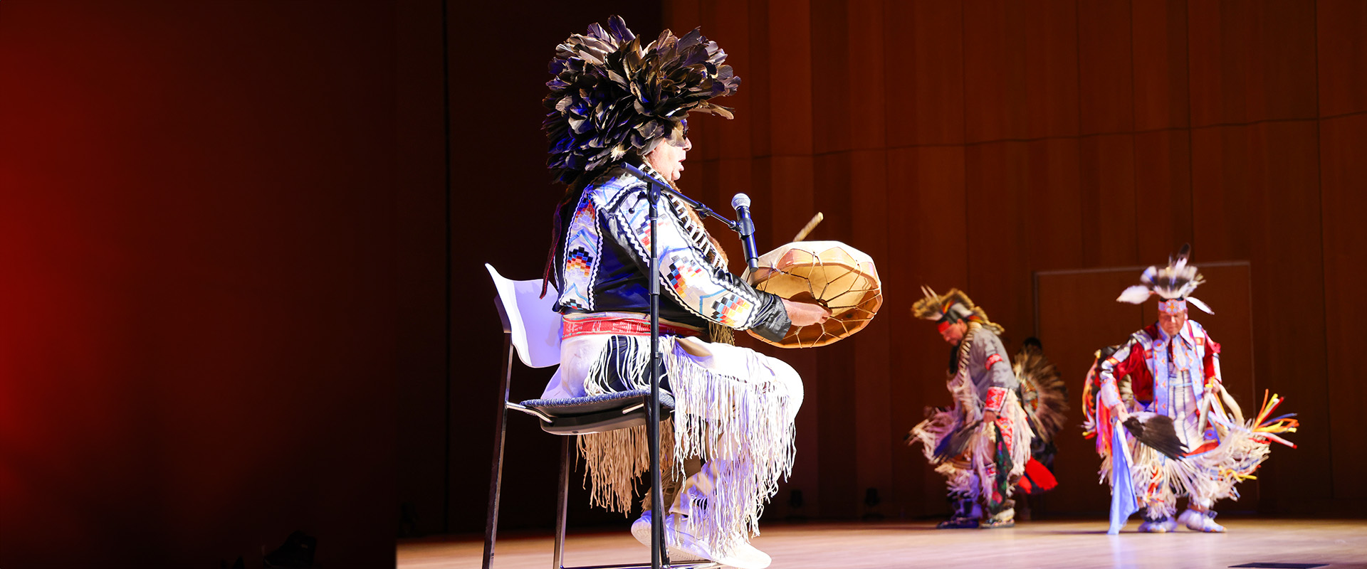A performer in traditional Indigenous attire plays a hand drum on stage while two others in regalia dance in the background.