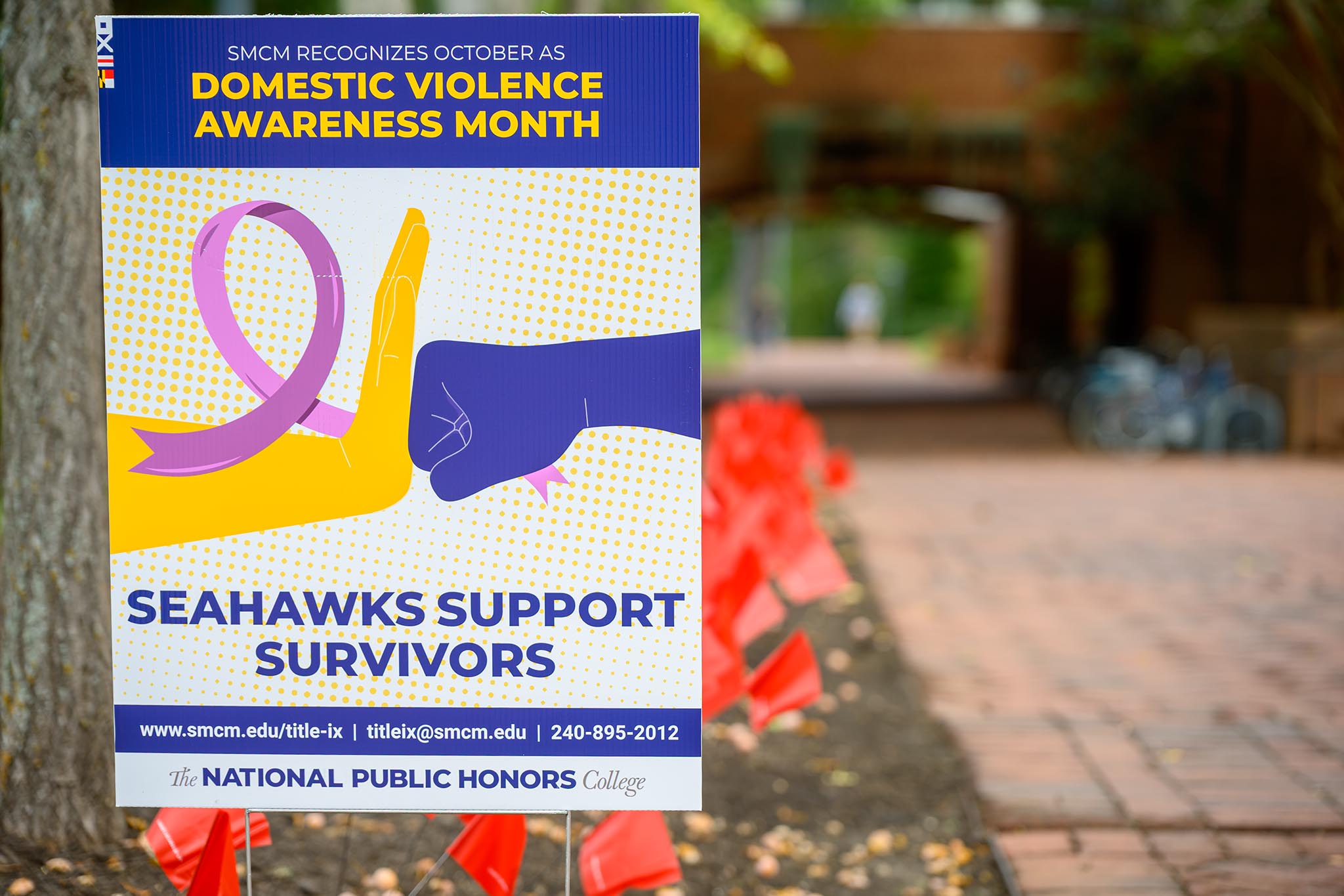 A sign on a brick walkway promotes Domestic Violence Awareness Month, featuring a purple ribbon and slogans supporting survivors, with red flags placed in the background.