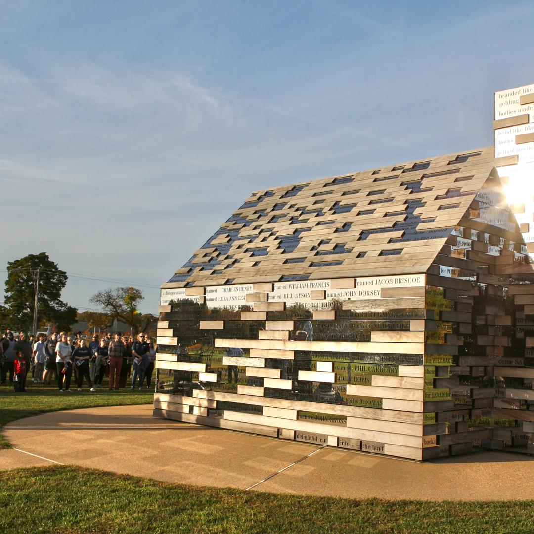 A group of people observe a large outdoor sculpture shaped like a house, constructed from rectangular reflective metal blocks, on a grassy field under a clear sky.