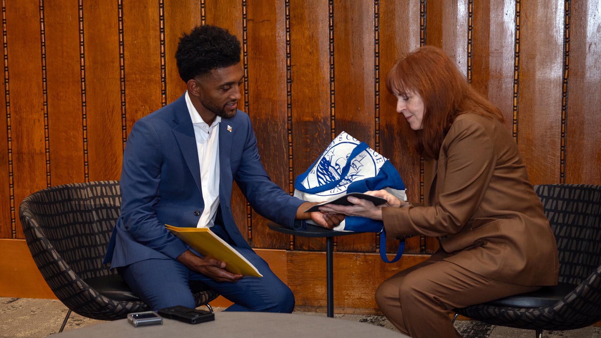A man in a blue suit and a woman in a brown suit sit in chairs by a small table, looking at a tablet together. Blue-and-white tote bag and documents are on the table.