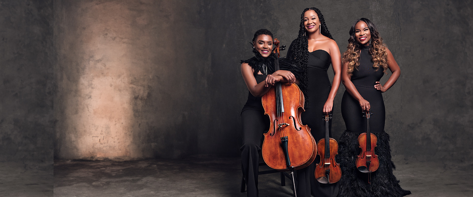 Three women in black evening gowns pose with string instruments, including a cello and two violins, against a neutral, textured backdrop.