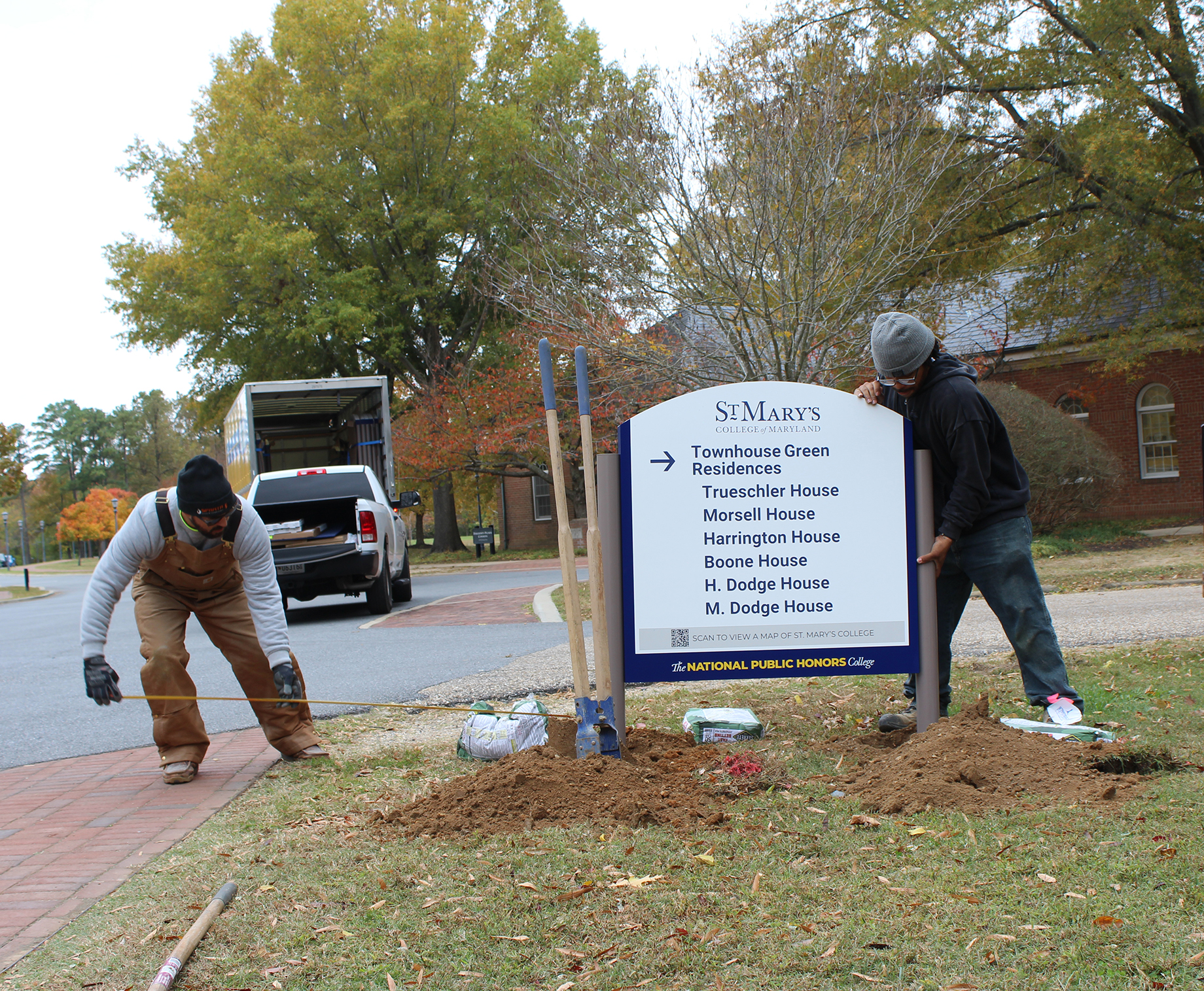Two workers install a St. Mary's College directional sign listing campus residences, with soil and tools on the ground nearby. A pickup truck is parked in the background.