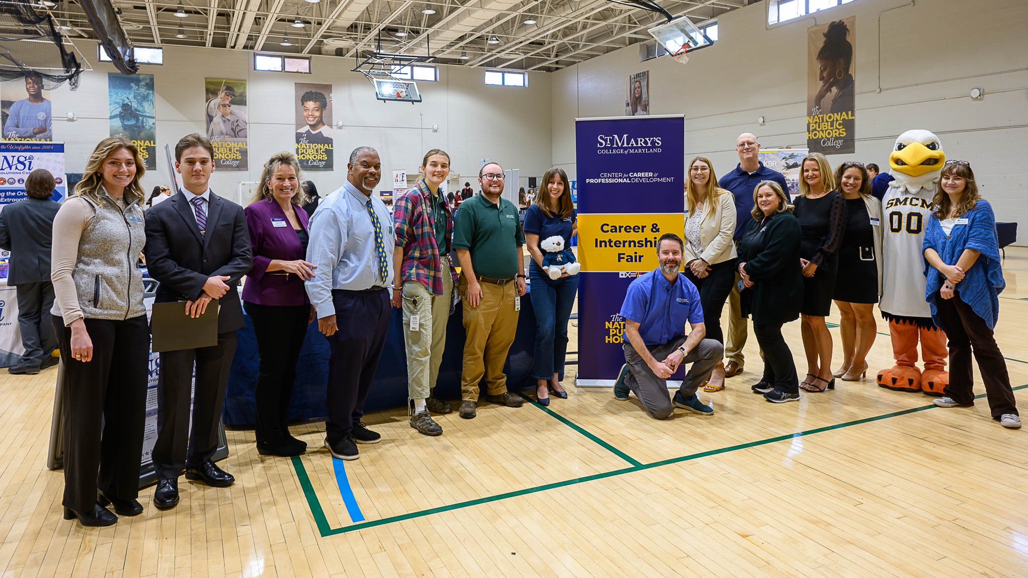 A group of people stands in a gymnasium next to a “St. Mary’s Career & Internship Fair” banner, with tables and a mascot visible in the background.