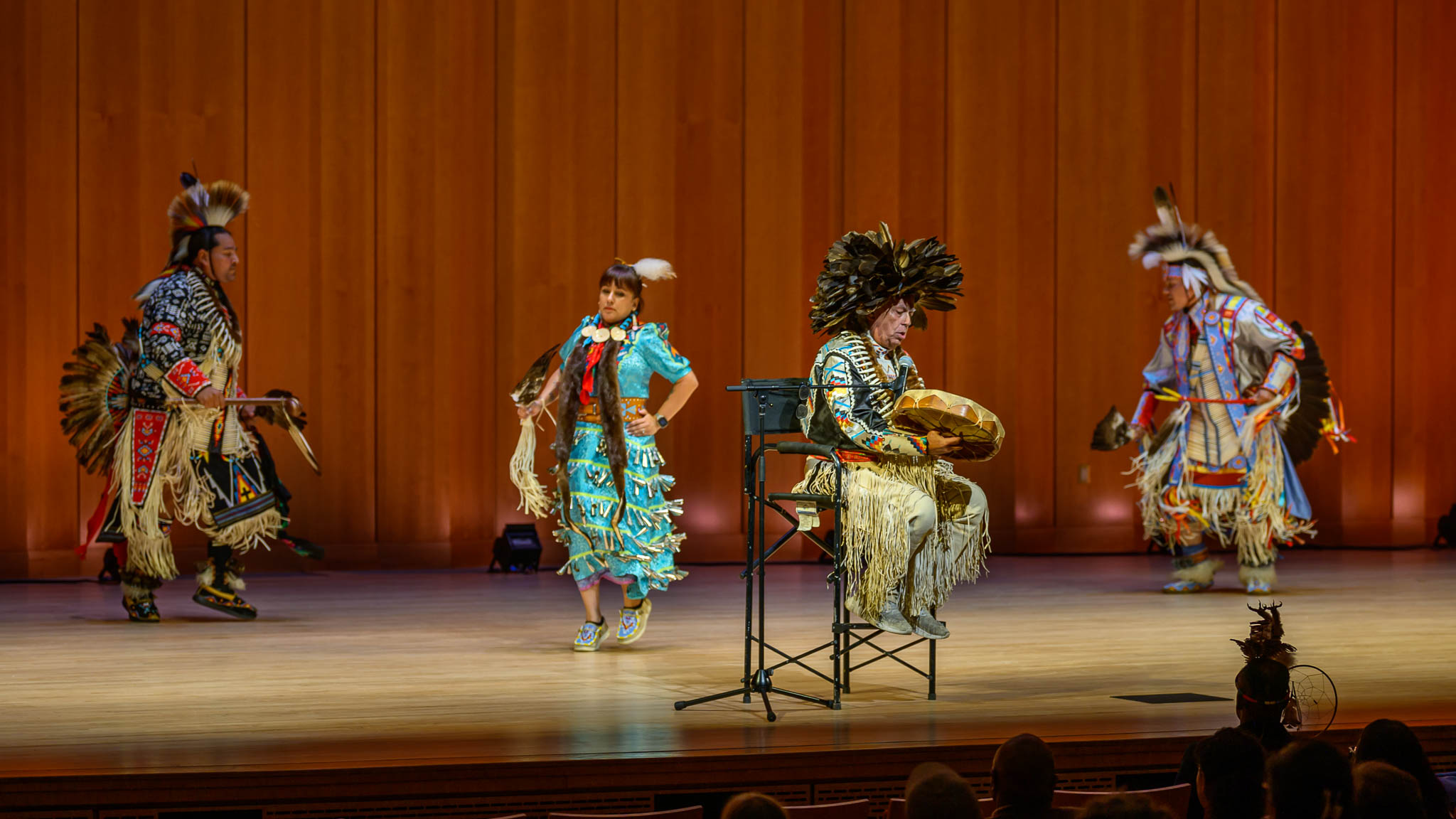 Four people in traditional Native American regalia perform on a wooden stage; one seated person plays a drum while three others dance in the background.