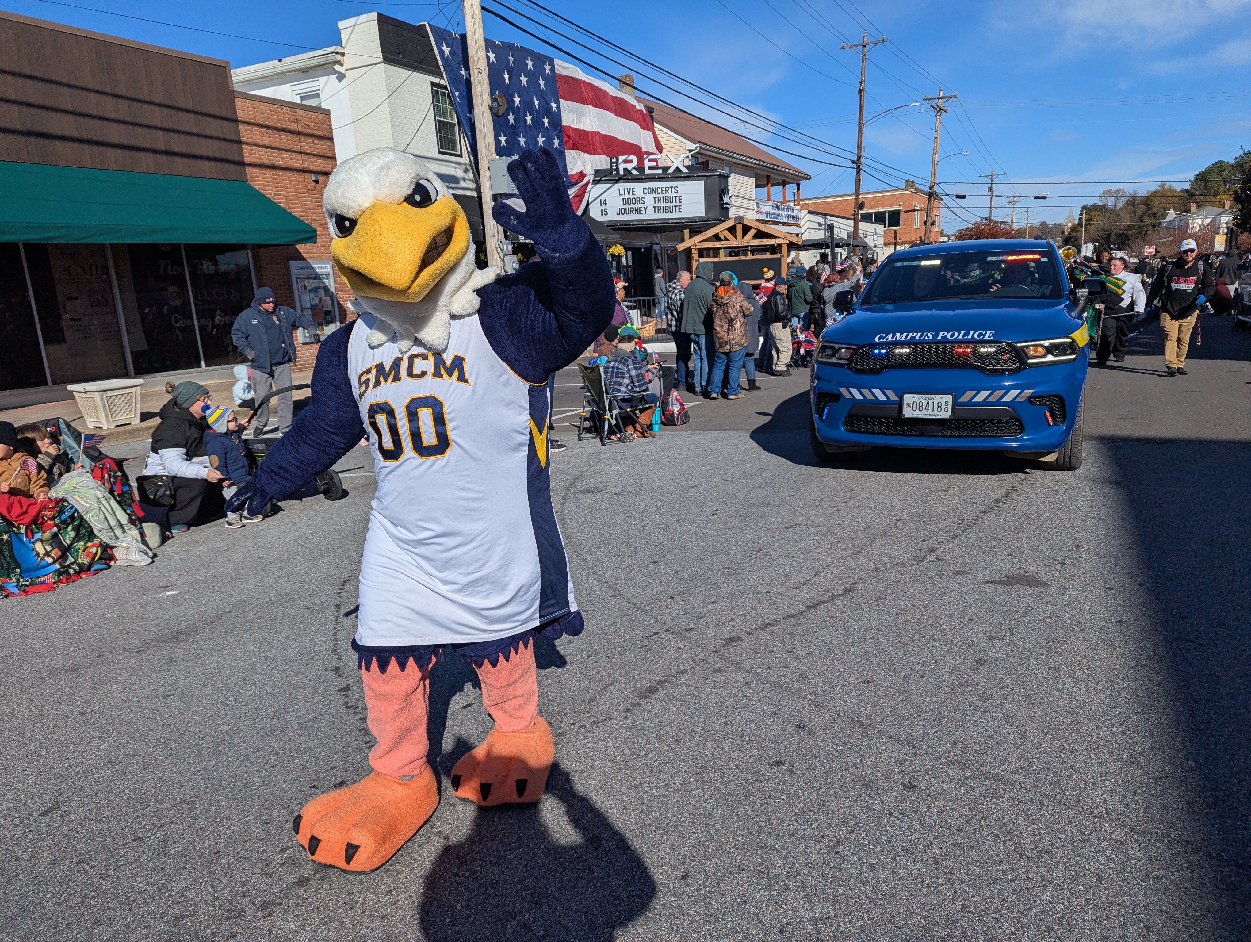A person in an eagle mascot costume wearing an SMCM jersey walks in a street parade next to a blue campus police car, with spectators on both sides.