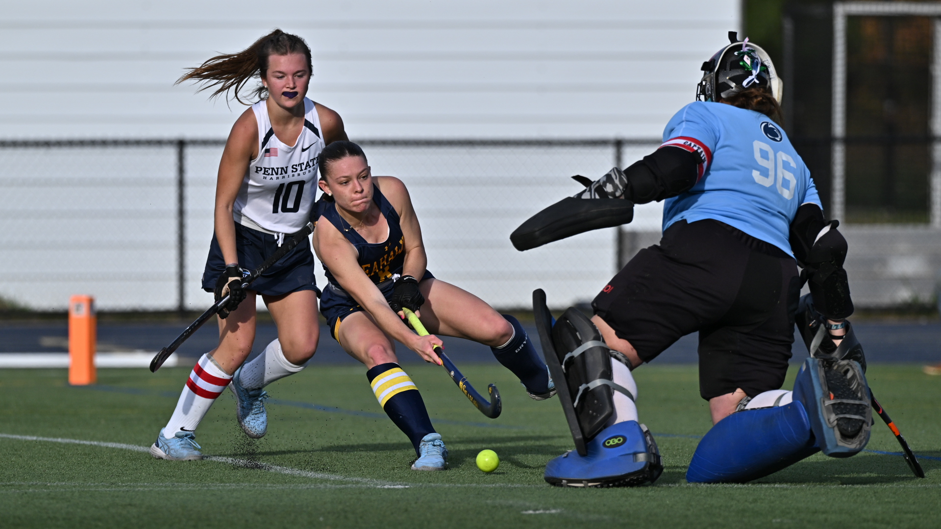 A field hockey player in blue attempts to hit the ball past a goalie in blue gear, while another player in white follows closely behind.