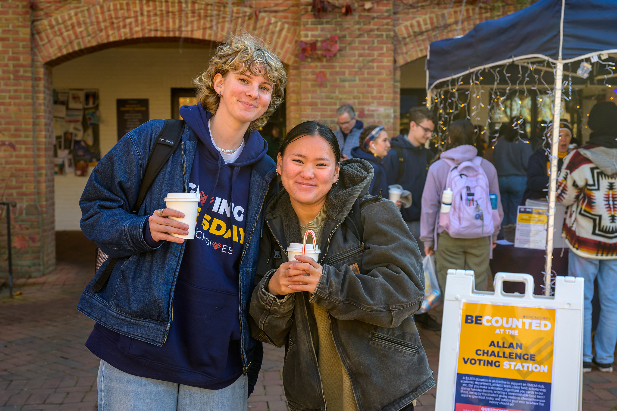 Two people stand outside near a voting station sign, holding drinks and smiling at the camera; other people can be seen in the background.