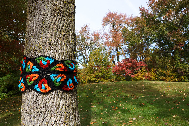A tree trunk wrapped with a colorful crocheted yarn pattern stands in a grassy park with autumn foliage in the background.
