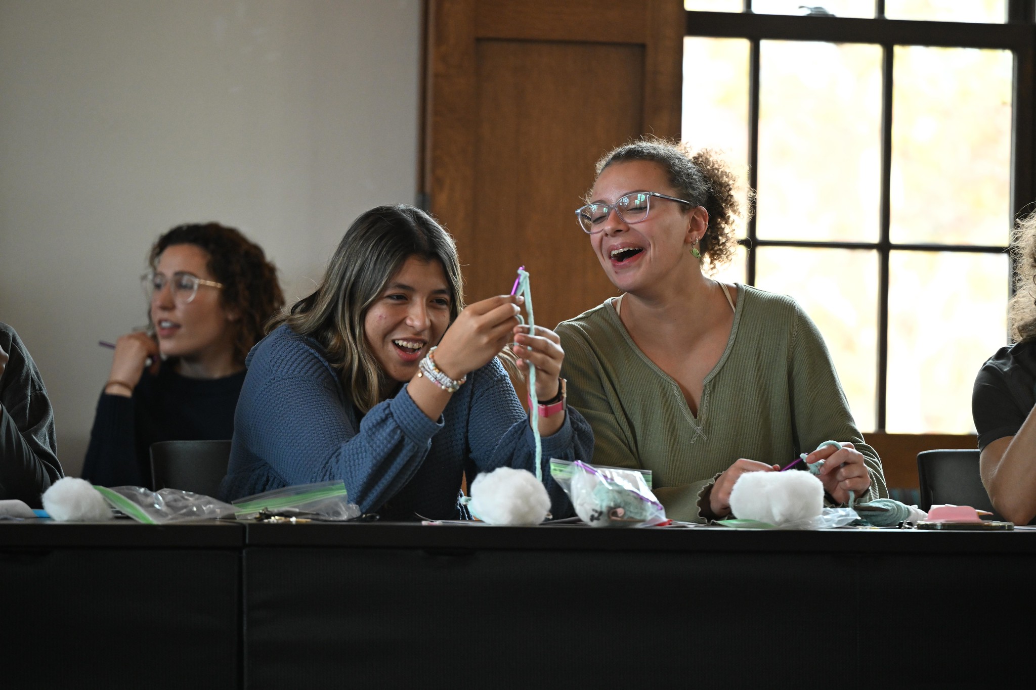 Two women sit at a table, smiling and laughing while holding craft supplies. Other people are seated nearby, and the table has bags of materials and cotton balls.
