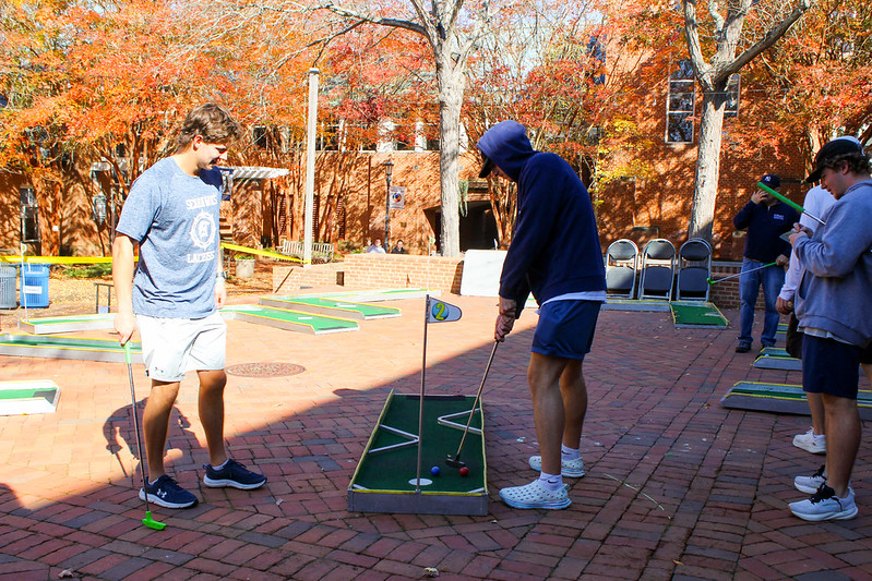 Several people play mini-golf on a brick courtyard surrounded by autumn trees and a brick building under sunny weather.