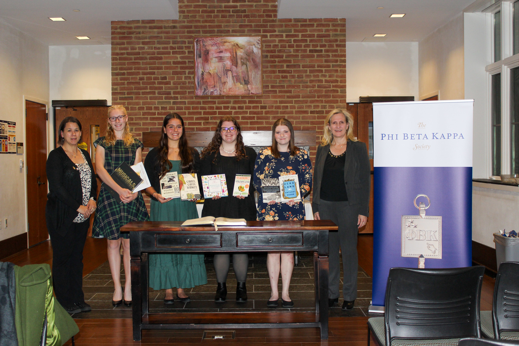 Six women stand indoors behind a desk with books, next to a "Phi Beta Kappa Society" banner. They are dressed formally and looking at the camera.